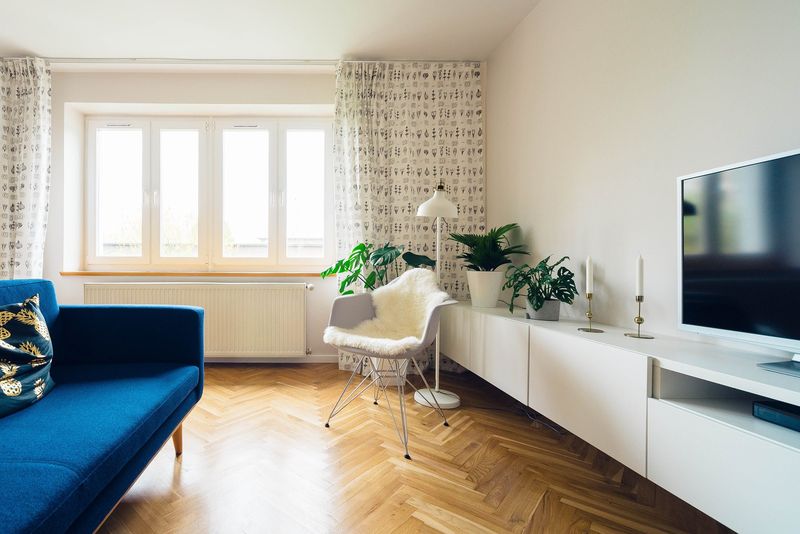 Salon lumineux avec une grande fenêtre blanche à double vitrage, un radiateur sous l'appui, un canapé bleu et un parquet à bâtons rompus