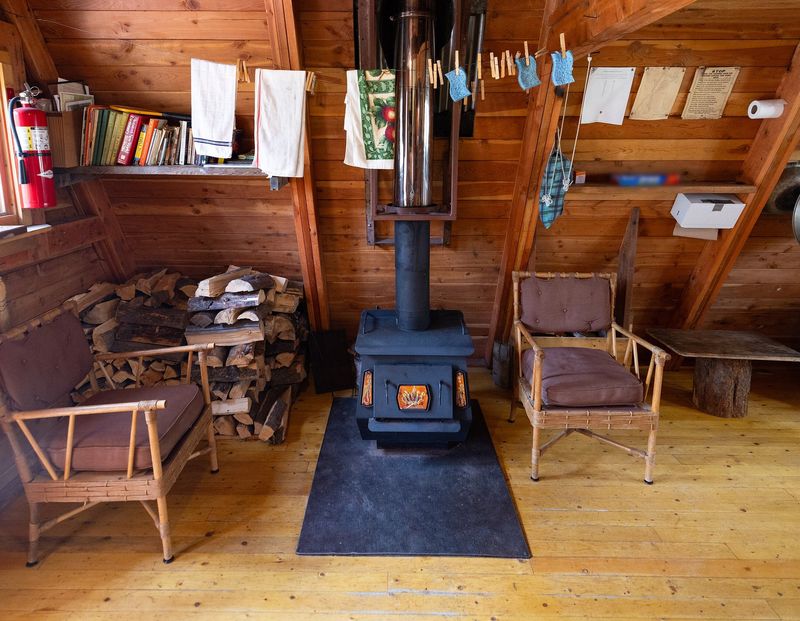Poêle à bois en fonte noire sur une plaque de sol, entre deux fauteuils, avec une réserve de bûches, dans un intérieur lambrissé de bois