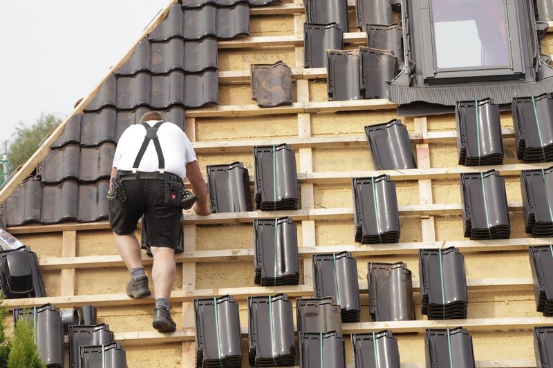 Couvreur de dos grimpant sur les liteaux d'un toit en réfection, piles de tuiles noires prêtes à poser et fenêtre de toit